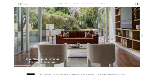 A modern living room featuring a brown sofa, two white armchairs, and a coffee table. Large windows open to a garden view. The room has wooden flooring and shelves with decorative items.