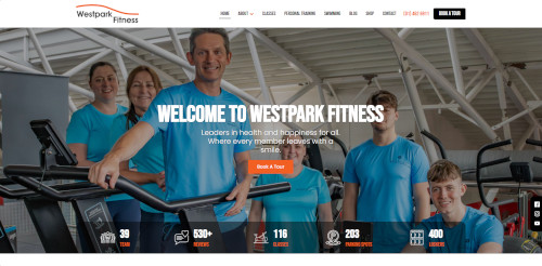 A group of fitness trainers wearing blue shirts pose together in a gym. They are smiling and standing near exercise equipment. The background features a spacious gym interior with bright lighting.